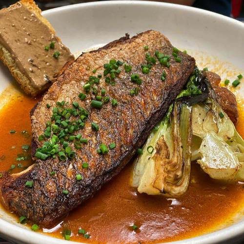A beautifully plated dish featuring seared fish, garnished with chives, alongside grilled vegetables and a slice of dessert.