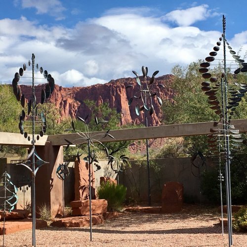 A desert landscape featuring unique metal sculptures against a backdrop of red rock formations and cloudy skies.