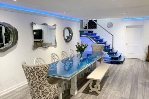 Modern dining area with a blue-tinted table, elegant chairs, mirrors, and a staircase with blue lighting.