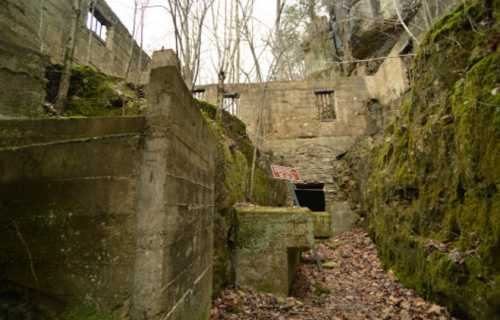 Abandoned concrete structure surrounded by moss-covered rocks and trees, with a sign partially visible in the background.