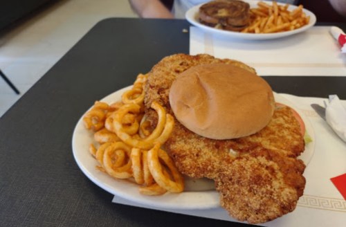 A plate with a large fried chicken fillet, a bun, and curly fries on a black table.