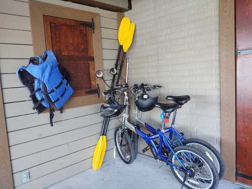 A blue life jacket hangs on a wall next to two bicycles and a pair of yellow kayak paddles.