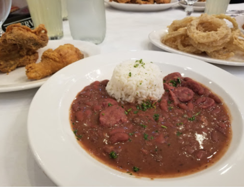 A plate of red beans and rice with sliced sausage, garnished with parsley, alongside fried chicken and onion rings.