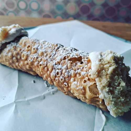 A close-up of a cannoli with powdered sugar on a piece of parchment paper, resting on a wooden surface.