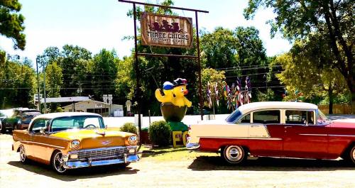 Two classic cars parked in front of a quirky sign featuring a large rubber duck and colorful decorations.