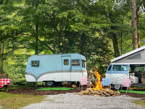 A vintage blue camper and a white van near a campfire, surrounded by trees and outdoor seating.