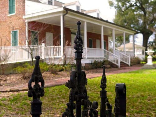 A historic brick house with white columns and a wrought iron fence in a landscaped yard.