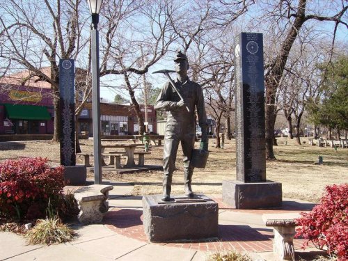 A bronze statue of a miner stands in a park, surrounded by stone pillars and trees, with benches nearby.