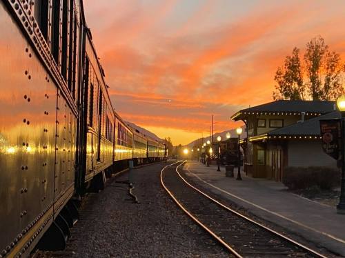A train on tracks at sunset, with vibrant orange skies and a station illuminated by warm lights.
