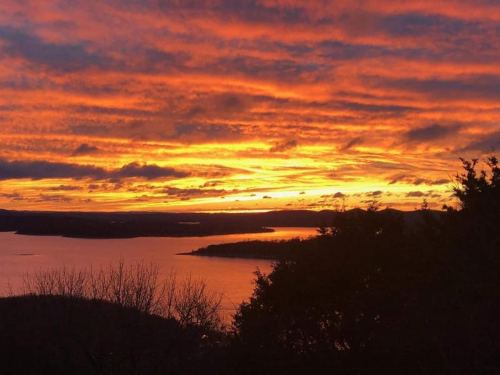 A vibrant sunset over a lake, with dramatic orange and purple clouds reflecting on the water. Silhouetted trees in the foreground.