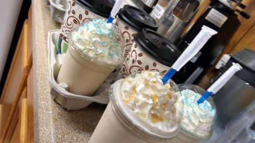 Four drinks with whipped cream and colorful toppings, placed in a drink carrier on a countertop.