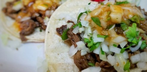 Two tacos filled with seasoned meat, topped with diced onions, cilantro, and salsa on a soft tortilla.