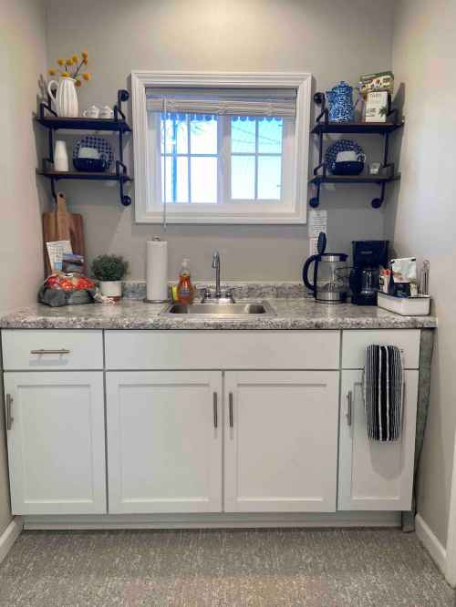 A small kitchen area with a sink, shelves, and coffee maker, featuring decorative items and a window above the sink.