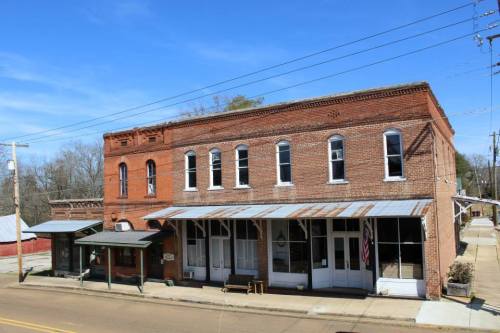 Historic brick buildings line a quiet street, featuring a covered porch and an American flag. Clear blue sky above.