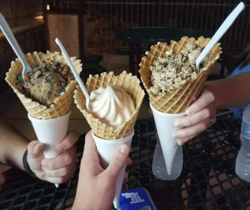 Three hands holding ice cream cones with different flavors and toppings, set against a dark outdoor background.