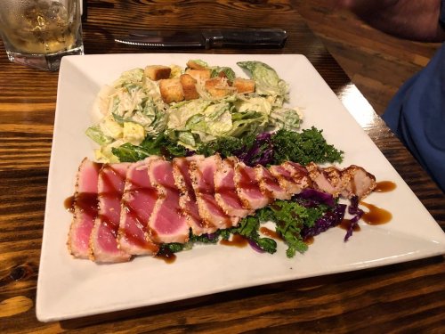 A plate featuring seared tuna slices, kale, purple cabbage, and a side Caesar salad with croutons.