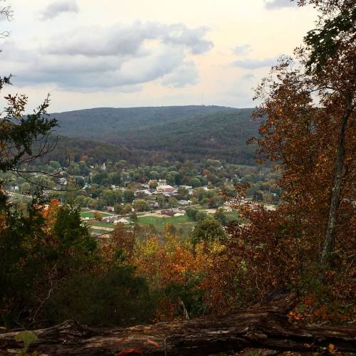 A scenic view of a small town surrounded by hills and trees, with a cloudy sky above.