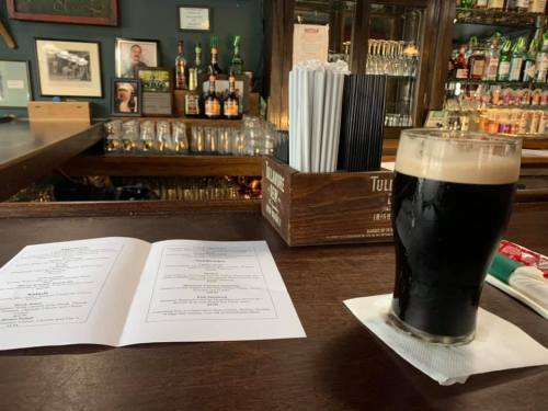 A pint of dark beer sits on a bar counter next to a menu, with shelves of liquor in the background.