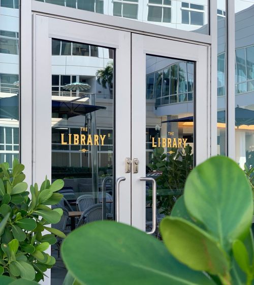 Glass doors with the text "THE LIBRARY" reflected, surrounded by green plants and a modern building backdrop.