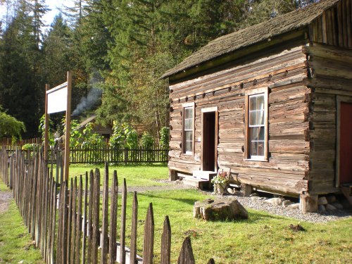A rustic wooden cabin surrounded by a picket fence and trees, with smoke rising from a nearby structure.