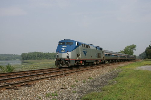An Amtrak train travels along a track beside a river, surrounded by greenery under a cloudy sky.