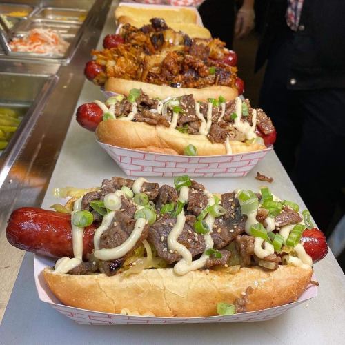 Three gourmet hot dogs topped with beef, onions, and sauces, served in buns on a counter.