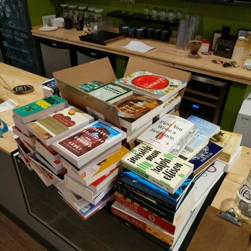 A stack of books on a wooden counter, with a box of books nearby and coffee equipment in the background.