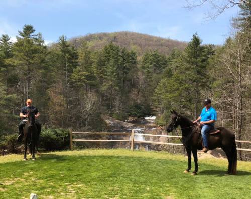 Two men on horseback in a grassy area, with a waterfall and trees in the background on a sunny day.