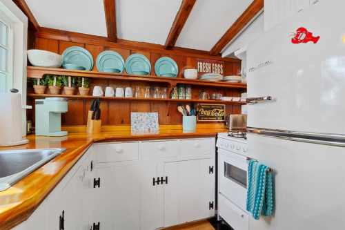 Cozy kitchen with wooden beams, white cabinets, vintage stove, and shelves displaying dishes and plants.