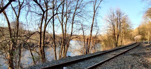 A serene view of a river alongside a railway track, framed by bare trees and a clear blue sky.
