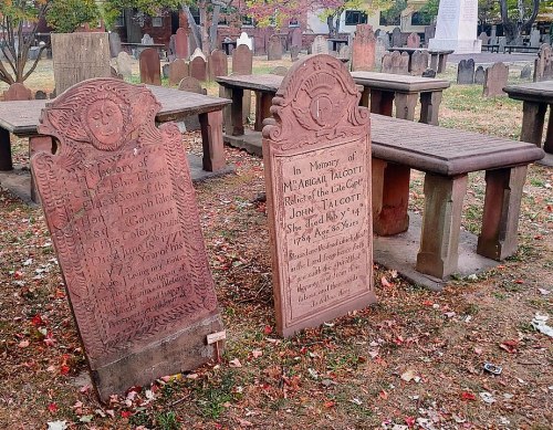 Two old gravestones in a cemetery, surrounded by weathered benches and autumn foliage.