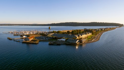 Aerial view of a marina with boats, waterfront buildings, and a distant shoreline under a clear blue sky.