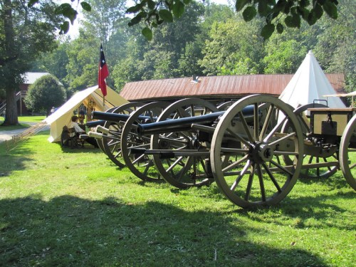 A row of historical cannons on a grassy field, with tents and a flag in the background.