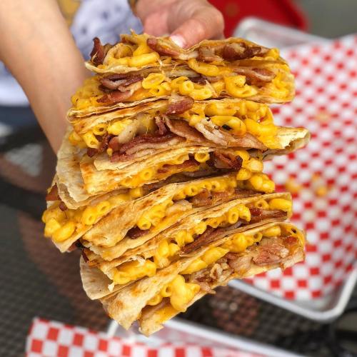 A hand holding a tall stack of cheesy bacon mac and cheese quesadillas on a checkered paper background.