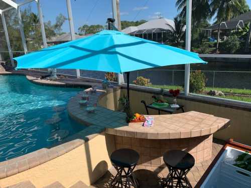 A bright blue umbrella shades a bar area by a pool, with two stools and a view of a canal and palm trees.