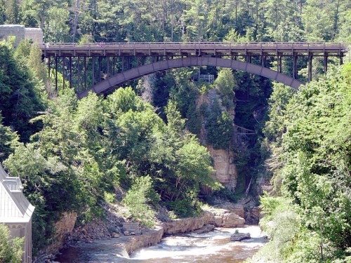 A metal arch bridge spans a river, surrounded by lush green trees and rocky cliffs.