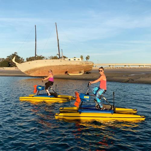 Two women paddle on yellow watercraft near a beached ship in a calm waterway under a clear sky.