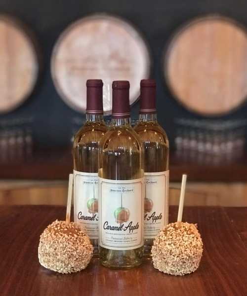 Three bottles of caramel apple wine with two caramel-coated apples on a wooden table, set against a backdrop of wine barrels.