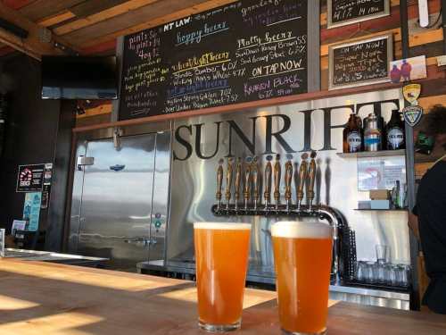 Two glasses of beer sit on a wooden bar in front of a colorful chalkboard menu at a brewery.