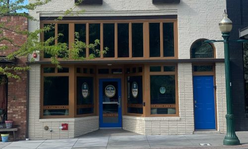 A storefront with large windows, blue door, and light-colored brick exterior, featuring a sign on the window.