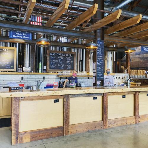 A rustic bar with wooden counters, chalkboard menus, and staff serving drinks in a spacious, industrial-style setting.