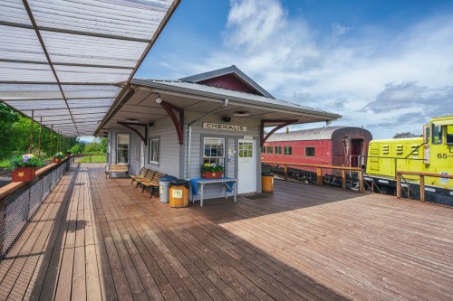 A wooden train station platform with benches, flower pots, and a yellow train car in the background under a blue sky.