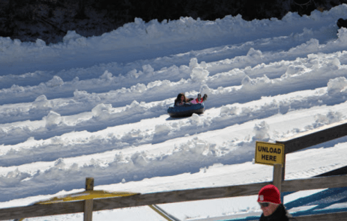 A child sleds down a snowy hill on a tube, with snow tracks visible and a sign that says "UNLOAD HERE" nearby.