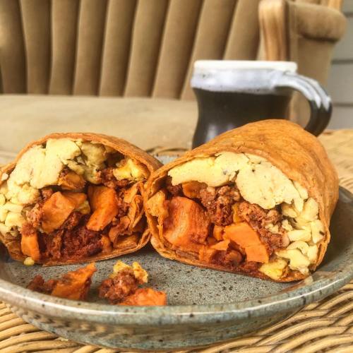 A plate with a cut burrito filled with sweet potatoes and scrambled eggs, next to a coffee mug on a wooden table.
