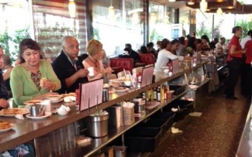 A busy diner with patrons seated at a long counter, enjoying meals and drinks in a lively atmosphere.