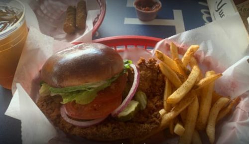 A crispy chicken sandwich with lettuce, tomato, and onion, served with fries and a drink in a red basket.