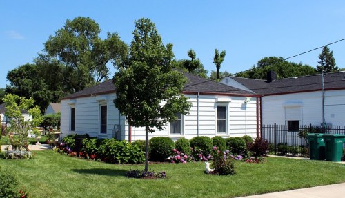 A white house with a black roof surrounded by green grass and colorful flower beds under a clear blue sky.