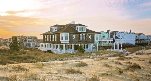 A large beach house with a sandy foreground, surrounded by smaller homes, under a colorful sunset sky.