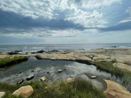 A serene coastal scene with rocky shores, calm water pools, and a cloudy sky reflecting over the ocean.