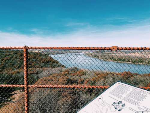 A view of a river surrounded by greenery, seen through a chain-link fence with an informational sign in the foreground.